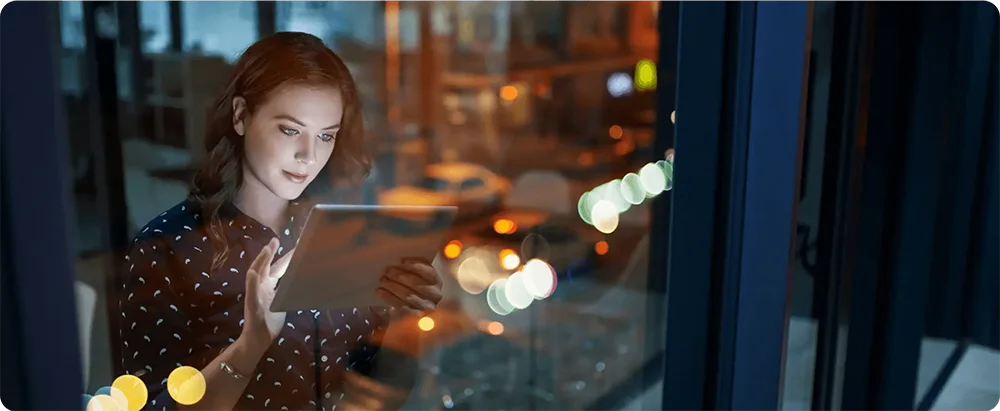 Woman using a tablet by the window with evening city lights.